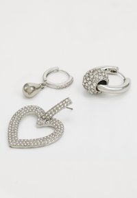 Zilveren oorbellen met sprankelende rhinestones: één hartvormig, één rond met een metalen ring, en een klein druppelontwerp, allemaal met glanzende afwerkingen.