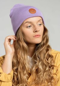 Fioletowa dziana czapka z prążkowanym wzorem i okrągłą brązową skórzaną naszywką z przodu. Nosi ją w połączeniu z żółtą koszulą z guzikiem.