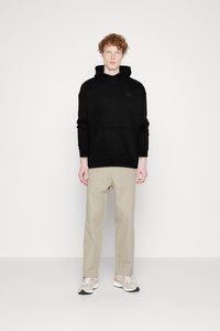 Felpa nera con tasca frontale e logo del marchio, abbinata a pantaloni beige chiaro e sneakers beige. Il modello è in piedi davanti a uno sfondo semplice.