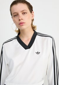 Fehér sportfelső fekete díszítéssel, valamint három fekete csíkkal, amelyek leérnek a hónaljig. A mellkasán egy kis fekete logó található.