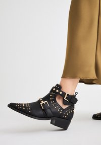 Botas negras hasta el tobillo de cuero con acentos de tachuelas doradas, hebillas y un tacón bajo. Los lados abiertos realzan el detalle visual.