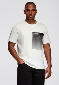 Bílý tričko s krátkým rukávem a černým proužkovaným designem na levé straně hrudi. Vyrobeno z měkké bavlny, s kulatým výstřihem.