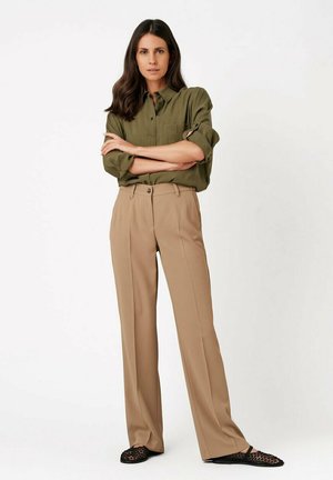 Frau mit langen dunklen Haaren, trägt ein olivgrünes Hemd mit Knöpfen und hochgekrempelten Ärmeln, beige weite Hosen und schwarze gewebte flache Schuhe, steht mit verschränkten Armen.