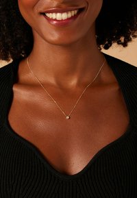 Gouden ketting met een kleine ronde hanger, tentoongesteld tegen een gestructureerd zwart kledingstuk. Hanger heeft een heldere, sprankelende detail.