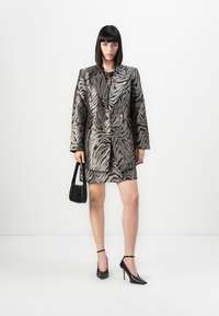 Kvinne i zebra-mønstrede blazer og skjørt, holder en liten svart håndveske, med svarte ankelrem-sandaler, stående på hvit bakgrunn.