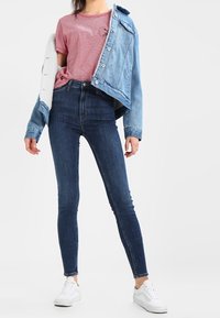 Chaqueta de mezclilla azul sobre una camiseta de rayas rojas, combinada con jeans ajustados oscuros y zapatillas blancas. Texturas suaves, ajuste casual.