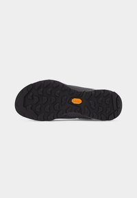Semelle de chaussure noire avec des crampons profonds et multidirectionnels et un logo Vibram orange au centre sur un fond gris clair uni.