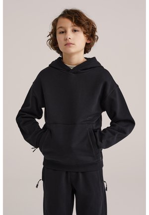 Copil cu păr creț, purtând un hanorac negru cu glugă și pantaloni asortați, stând cu mâinile în buzunare pe un fundal neutru.