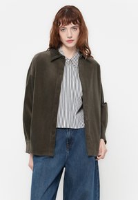 Tmavě zelená manšestrová oversized košile s límečkem, přepnutá přes černobílou pruhovanou košili s knoflíky. Volné džíny doplňují celý outfit.