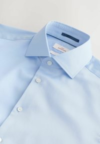 Chemise bleu clair à boutons en tissu lisse, avec un col italien et trois boutons à l'avant. Étiquettes visibles sur le col.