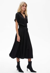 Zwarte wikkeljurk met korte mouwen, een versmalde taille en een vloeiende rok. Draagt een paar zwarte enkellaarsjes met een slank ontwerp en een spitse neus.