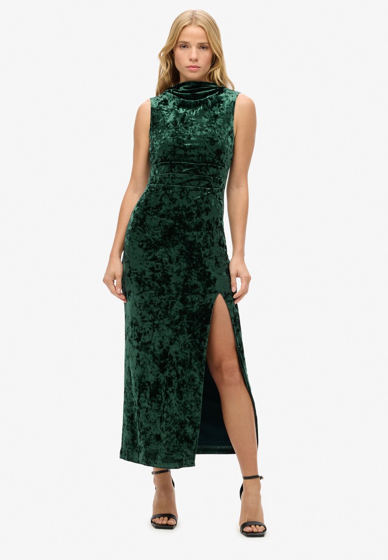 Robe maxi en velours vert avec un corsage ajusté, un col montant et une fente haute sur la cuisse. Texture lisse et design épuré ; portée avec des sandales noires à lanières.