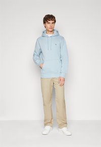 Light blue hooded sweatshirt with front kangaroo pocket, drawstrings, and subtle logo on the chest, paired with beige trousers and white shoes.