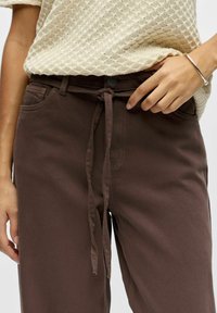 Pantalon en coton marron avec taille à cordelette, texture lisse, nervures diagonales subtiles et détails de coutures jaunes contrastantes aux poches.