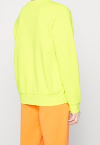 Neon žlutá mikina s dlouhými rukávy a žebrovanými manžetami, spárovaná s oranžovými kalhotami, předvádějící uvolněný střih a hladkou texturu.