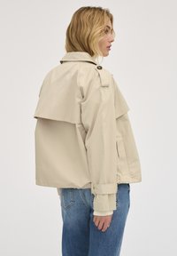 Beige zkrácená bunda s vrstveným designem na zádech, velkým límcem a knoflíkovými detaily. Má široké rukávy a volný střih.
