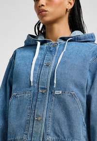 Vaaleansininen farkkutakki, jossa on huppu, edessä napit ja kaksi suurta taskua. Valkoiset narut ja pieni logopatchi.