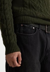Maglione in maglia a treccia verde scuro con polsini a coste, abbinato a jeans di denim neri con una cintura nera e bottoni rotondi. Ingrandimento della mano.