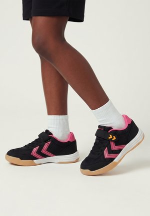 Ben med sorte og pink atletiksneakers og hvide sokker, stående mod en ensfarvet lys baggrund.