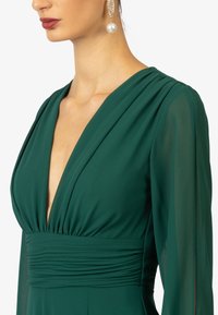 Groene jurk met een diepe V-hals, geplooide lijfje en lange, doorzichtige mouwen. Opmerkelijke details zijn de elegante stoftextuur en draping.