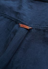 Tissu bleu marine avec un pli central et une petite étiquette de marque orange "SUPERDRY" cousue le long de la couture.