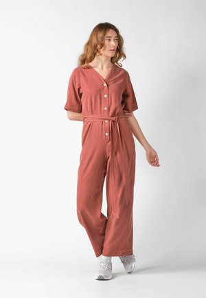 Woman with wavy hair wearing rust-colored buttoned jumpsuit with belt, standing and looking sideways, white sneakers on plain background.