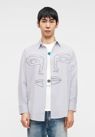 Ριγέ φωτεινό γαλάζιο πουκάμισο με αφηρημένο σχέδιο προσώπου σε μαύρο περίγραμμα, με μακριά μανίκια και κλείσιμο με κουμπιά, φορεμένο πάνω από λευκό T-shirt.