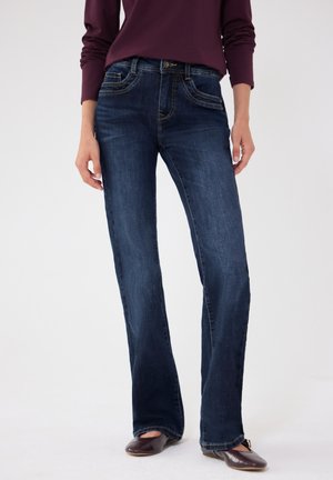 Kvinde iført mørkeblå bootcut-jeans, mørkelilla langærmet top og brune flade sko, stående mod en ensfarvet lys baggrund.