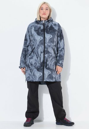 Persona vestita con una giacca con cappuccio blu-grigio marmorizzata con tasche frontali, pantaloni neri e scarpe da trekking scure con lacci rossi, in piedi contro una parete bianca.