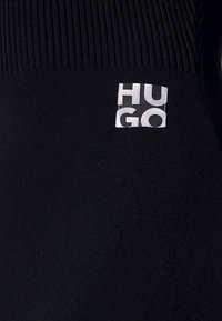 Zwarte gebreide stof met een ribbelige bovenkant. Aan de linkerkant is een wit label met "HUGO" in vette zwarte letters gestikt.