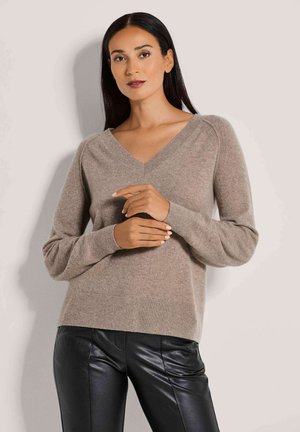 Beiger V-Ausschnitt-Pullover aus weichem Material mit langen Ärmeln, gerippten Bündchen und lockerer Passform, kombiniert mit schwarzen Lederhosen.