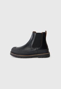 Bottines Chelsea en cuir noir avec semelle épaisse, panneaux latéraux élastiques et tirant marron sur fond gris clair uni.
