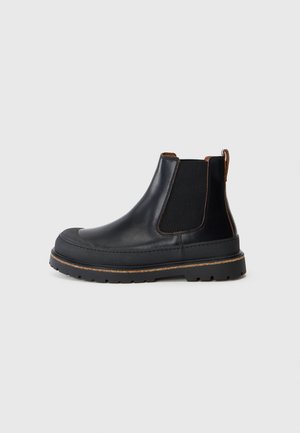 Botín Chelsea de cuero negro con suela gruesa, paneles laterales elásticos y tira de agarre marrón sobre un fondo gris claro liso.