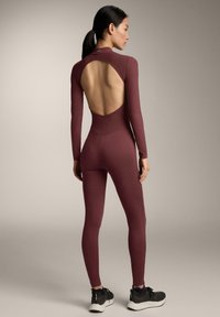 Bordó, teljes testet borító overál hosszú ujjakkal, nyitott hátú kialakítással, bordázott textúrával és testhezálló sziluettel, fekete sportcipővel párosítva.