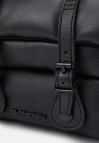 Primo piano di una borsa in pelle nera con cinturino a fibbia cucito e logo Dr. Martens impresso sulla superficie imbottita.