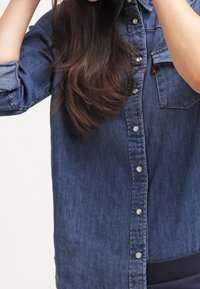 Chemise en denim bleu moyen, à manches longues, avec un col classique et des boutons-pression argentés sur le devant. Dispose d'une poche poitrine.