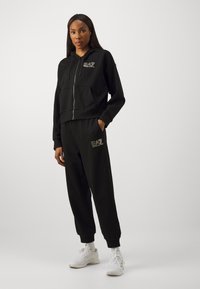 Črna zip-up kapuca z vpoglednimi žepi in usklajenimi hlačami, oba z zlatim logotipom EA7. Mehka, strukturirana tkanina z rebrastimi manšetami. Bele superge.
