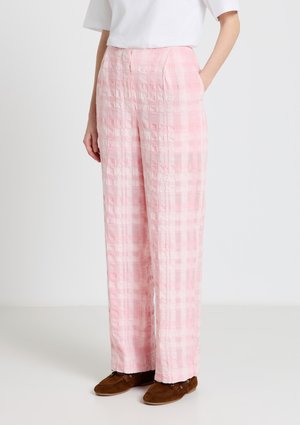 Personne portant un pantalon à taille haute, jambes larges, aux carreaux roses et blancs avec poches latérales, un haut blanc et des mocassins marron, debout sur un sol uni.