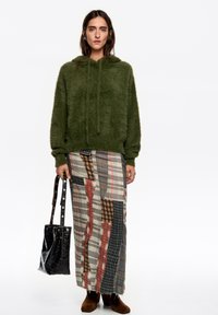 Sudadera con capucha verde y pelo suave, con cordones, combinada con una falda larga con múltiples patrones en diseños de cuadros; la modelo sostiene un bolso negro con textura.