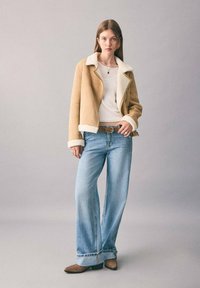 Beige Schaffelljacke mit weißer Fütterung, getragen über einem weißen Oberteil, kombiniert mit hellblauen weiten Jeans und braunen Ankle Boots.
