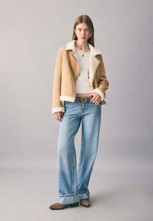 Veste en shearling beige avec doublure blanche, portée sur un top blanc, assortie à un pantalon en jean ample bleu clair et des bottes chelsea marron.