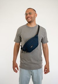 Marsupio di tessuto blu navy indossato a tracolla, dotato di due tasche con zip e una cintura nera regolabile. Texture liscia, design minimalista.
