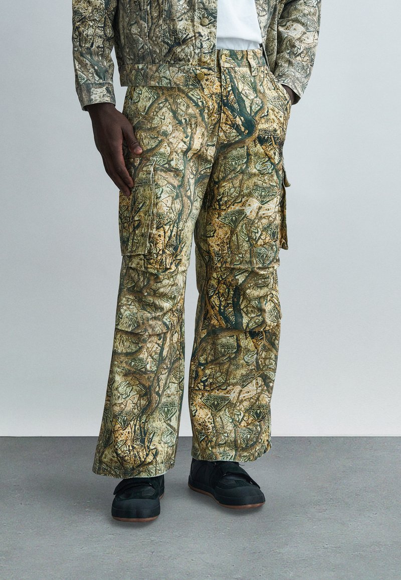 Παντελόνια καμουφλάζ cargo σε πράσινους, καφέ και κίτρινους τόνους· διαθέτουν τσέπες και άνετη εφαρμογή. Συνδυάζονται με μαύρα αθλητικά παπούτσια για αντίθεση.