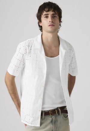 Junger Mann trägt ein weißes, kurzärmeliges Hemd mit Ösen über einem weißen Tanktop, beige Hosen und einen geflochtenen braunen Gürtel, steht vor einem einfarbigen Hintergrund.