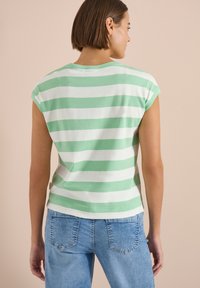 Femme aux cheveux courts et bruns, portant un haut sans manches rayé vert et blanc et un jean bleu clair, de dos sur un fond beige.