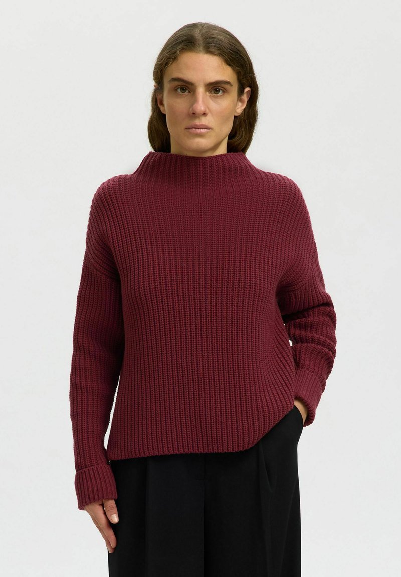 Femme portant un pull en maille épaisse bordeaux à col montant et un pantalon noir, debout avec une main dans la poche.