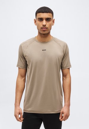 Jaunuolis trumpais plaukais dėvi smėlio spalvos BOSS trumpomis rankovėmis marškinį ir juodus kelnes, stovėdamas priešais paprastą šviesią foną.