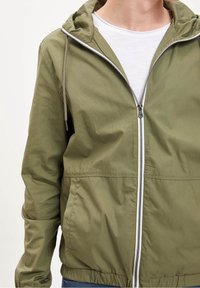 Olivgrüne Zip-Jacke mit Kapuze, elastischen Bündchen und Saum. Verfügt über einen grauen Reißverschluss und einen leichten, leicht strukturierten Stoff.