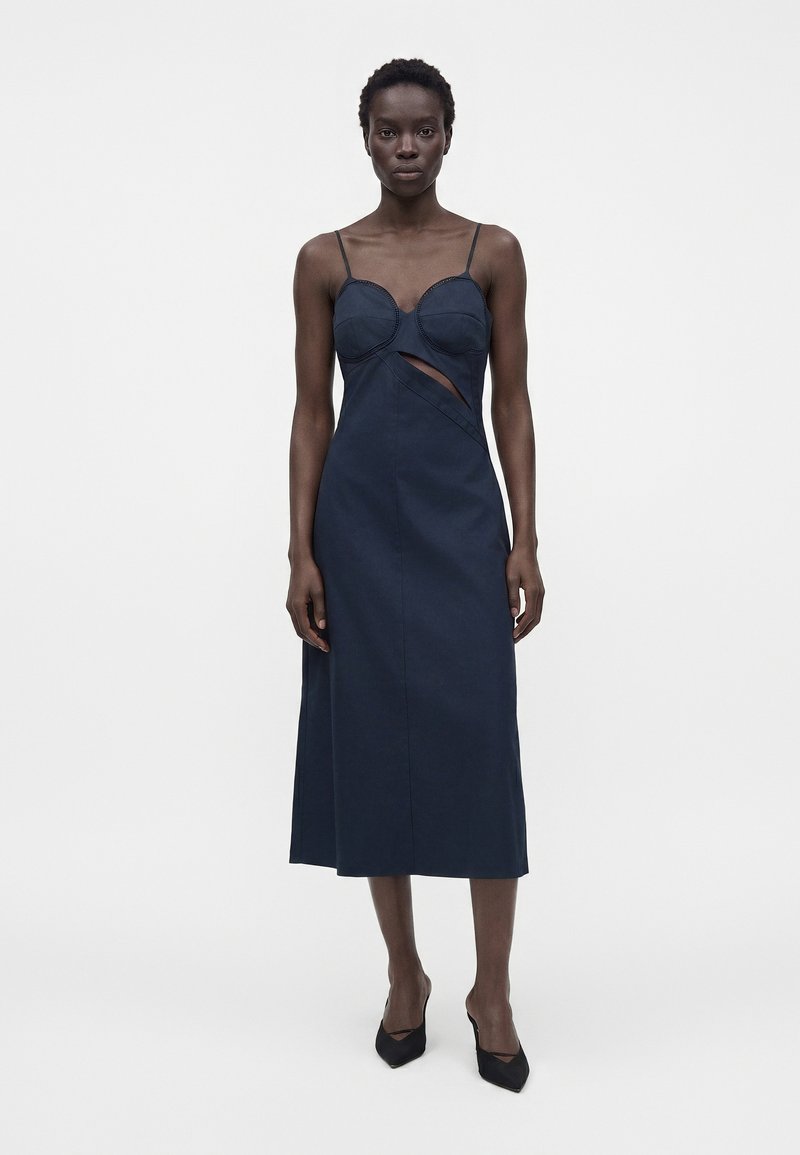 Woman with short hair wearing a navy blue mid-length dress with thin straps, a cut-out detail, and black pointed heels standing against a plain background.