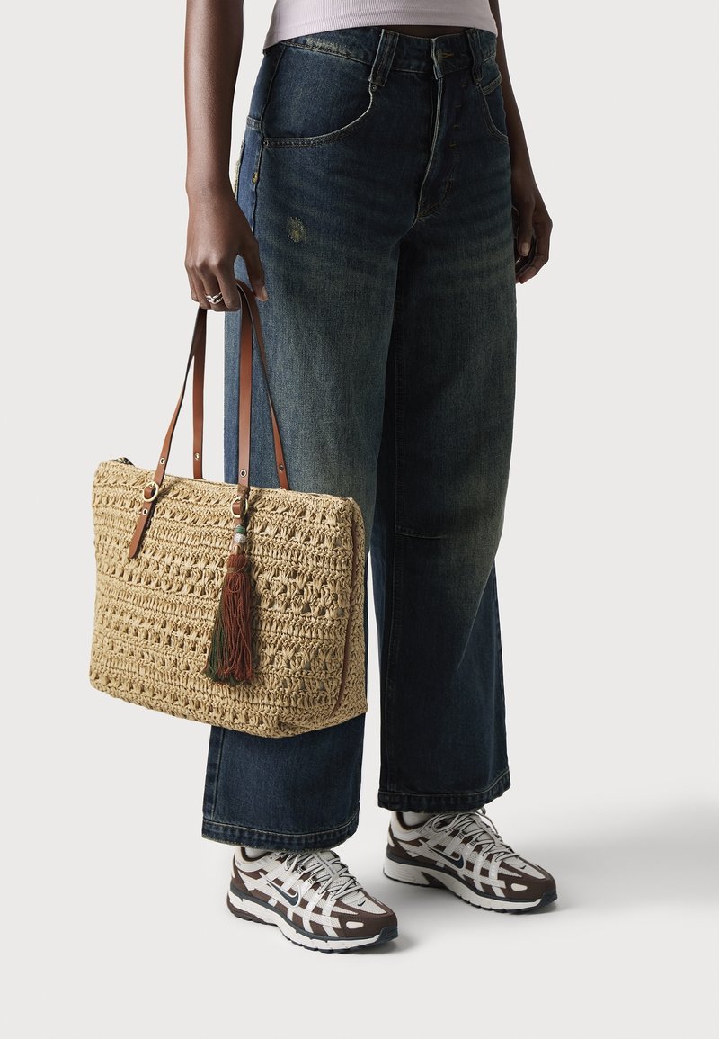 Personne portant un jean à jambes larges et des baskets, tenant un sac à main beige tressé avec des liens en cuir et un détail pompon.